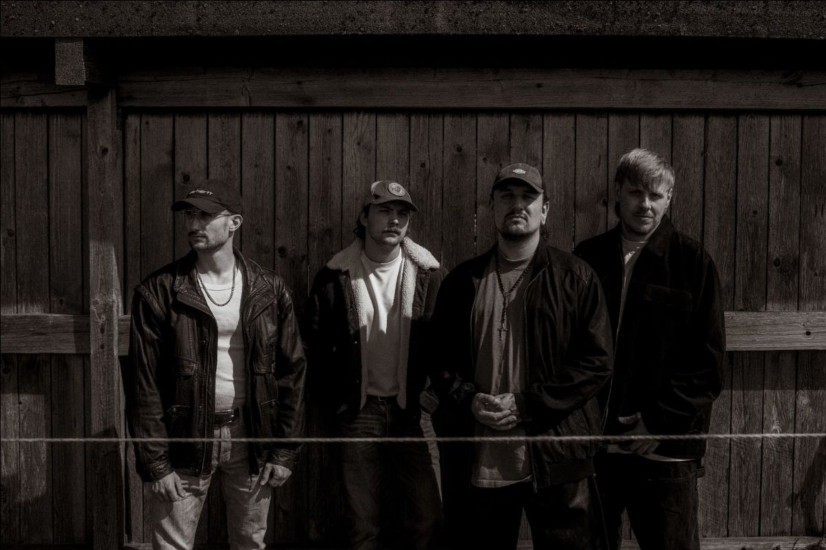 Photo de groupe en noir et blanc devant une palissade en bois, quatre musiciens en blouson