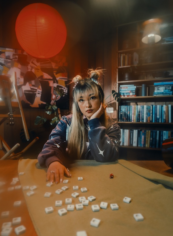 Portrait de RINRIN assise à une table, entourée de tuiles de jeu et d'une bibliothèque, sous une lanterne rouge