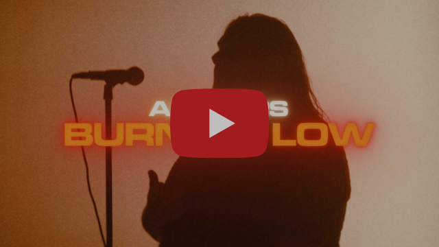 Silhouette d'un chanteur devant un micro, sur fond orangé, avec le titre Burning Low en lettres de néon