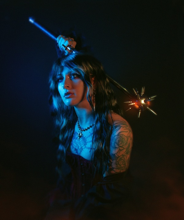 Portrait de Mothica en lumière bleue, tenant une tige lumineuse, avec des tatouages sur le bras