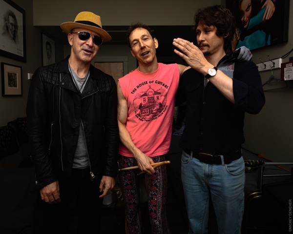 Trois hommes posant ensemble dans une loge, l'un en chapeau et veste en cuir, un autre en débardeur rose tenant des baguettes, le troisième en chemise sombre