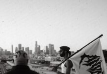 Ando San dévoile « March For Me », entre hip-hop et prog-metal Deux hommes sur un toit de Los Angeles, l'un tenant un drapeau orné d'un motif noir, avec la skyline de la ville en arrière-plan