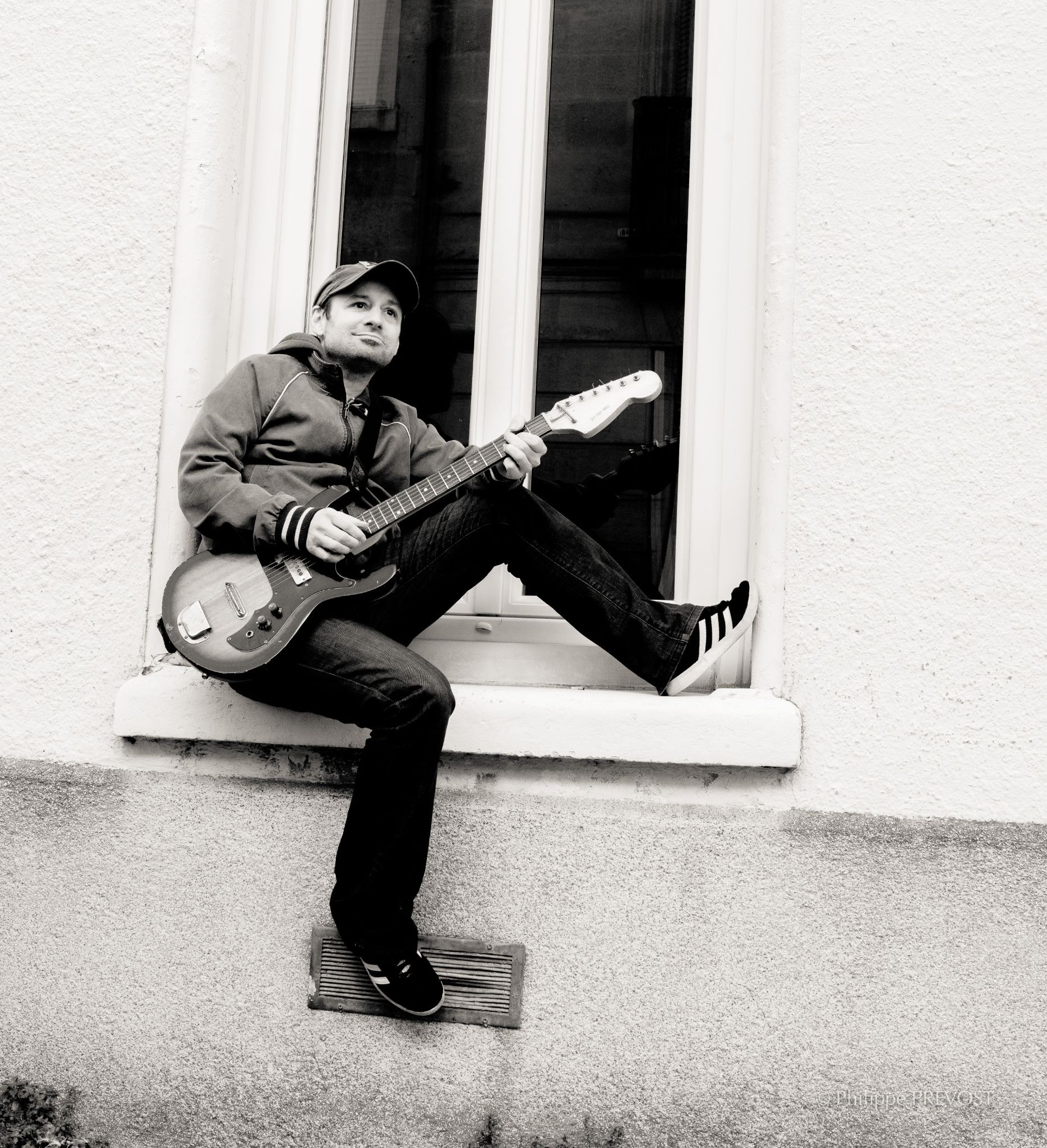Julien Pras assis sur un rebord de fenêtre, guitare électrique à la main, en noir et blanc
