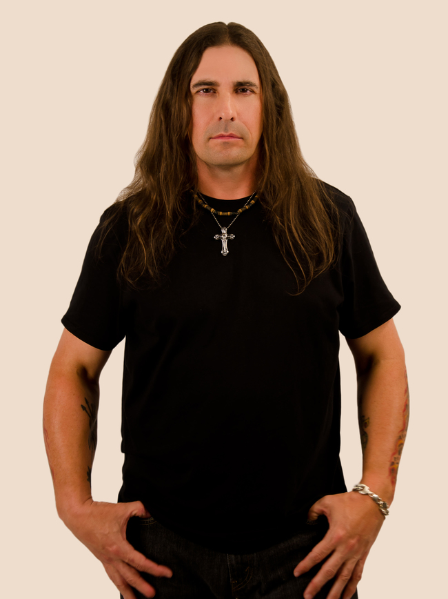 Portrait d'un homme aux cheveux longs, vêtu d'un t-shirt noir et d'une croix en pendentif, sur fond beige