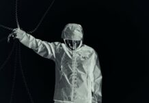 SUNBORN dévoile « Silent Attraction », un single au groove rugueux Silhouette en vêtements clairs et capuche, bras tendu vers des chaînes, sur fond noir